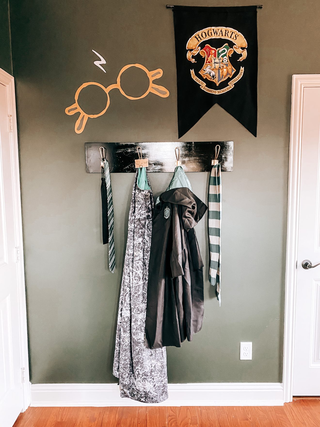 Wall decorated with Harry Potter Glasses decal, Hogwarts crest banner flag and wall hooks that have a Slytherin tie, Slytherin robe, Slytherin scarf and the cloak of invisibility hanging from it | The Yellow Rose Farmhouse | www.theyellowrosefarmhouse.com