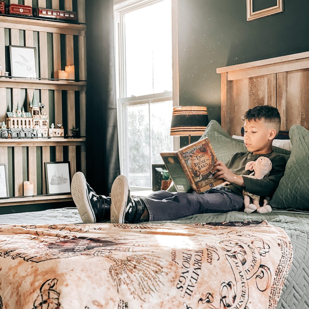 Boy reading Harry Potter on his Slytherin bed with the marauder's map throw blanket, a DIY wood slat wall with shelves that has hogwarts legos and the Hogwarts Express | The Yellow Rose Farmhouse | www.theyellowrosefarmhouse.com
